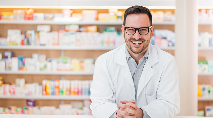 Zu sehen ist ein Apotheker im weißen Kittel und Schaugläsern vor Regalen voller pharmazeutischer Produkte.